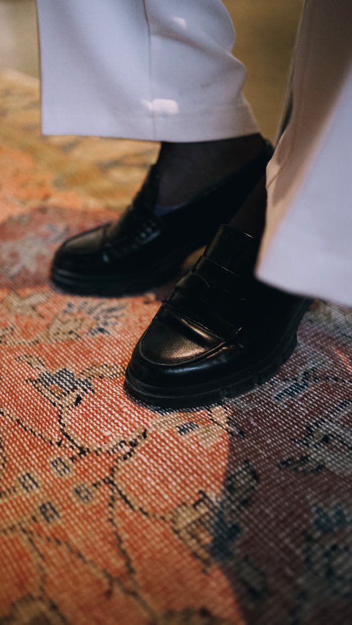 category-01 Close-up of black leather shoes and white trousers on a patterned carpet.